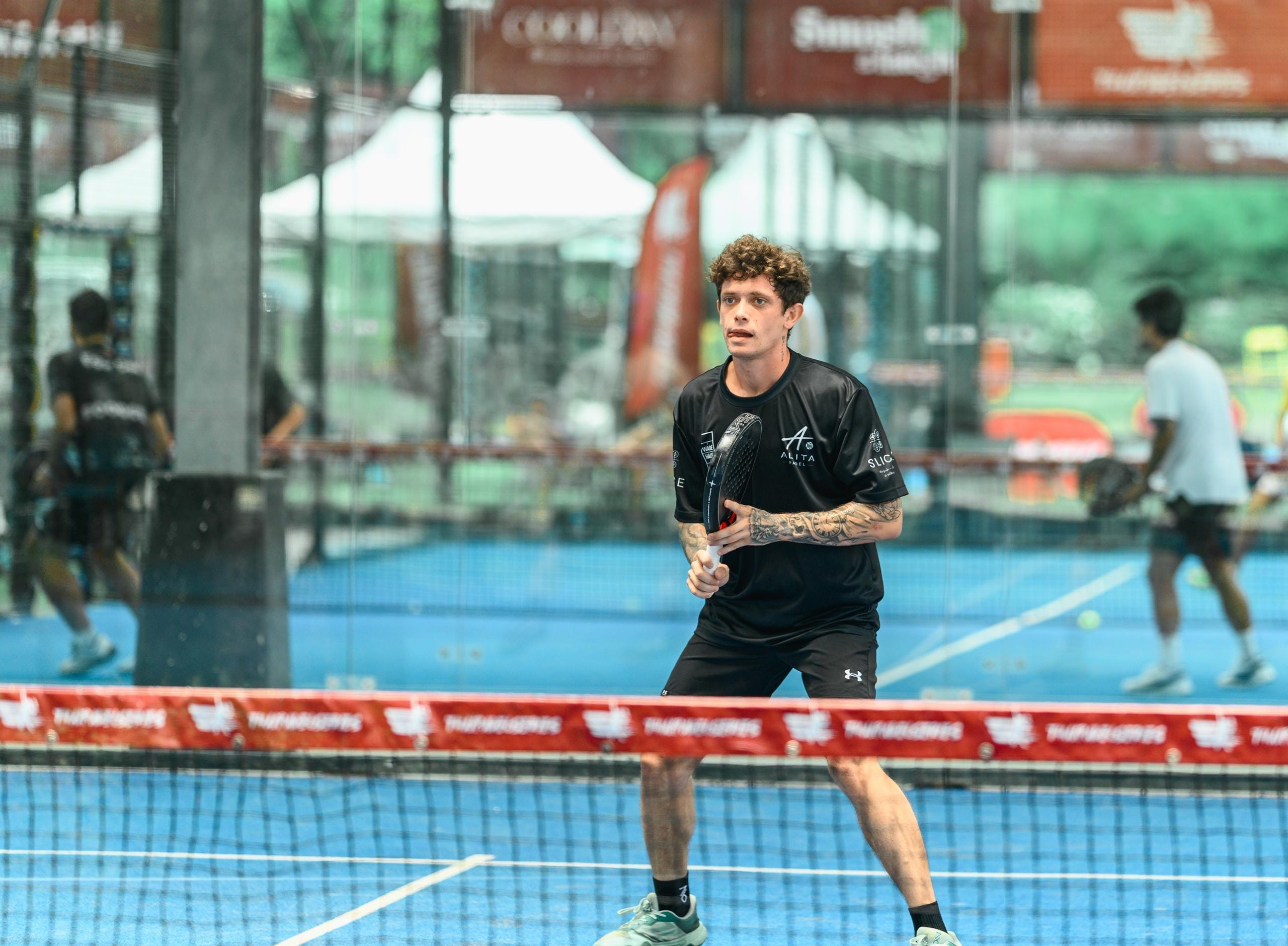 Coach Benji in a ready position at the net during a tournament match, black kit, blue court.