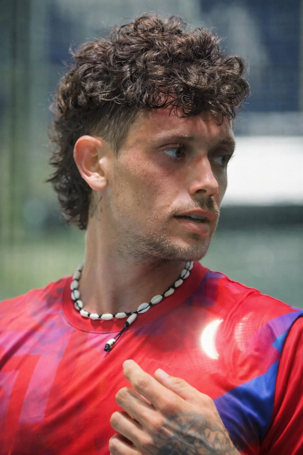 Close-up portrait of Coach Benji in a red shirt wearing a shell necklace, looking off court.